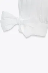 Camisa blanca de manga larga con cuello solapa y cierre frontal de botones. Los puños terminan con lazos anudados en el mismo tejido.