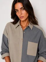 Camisa oversize de diseño bicolor, combinando paneles en color violeta y gris. Presenta cuello clásico, cierre frontal con botones, bolsillo en el pecho y mangas largas con puños abotonados.