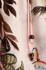 Vestido midi oversized de mezcla de viscosa, con estampado floreado tropical sobre base rosada, escote en V y botonadura frontal. Cuenta con mangas al codo raglán con puños fruncidos, cinturón fino para atar con cuentas y diseño escalonado en la falda.