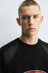 Camiseta negra de corte boxy fit con paneles laterales y de hombros en tejido de malla. Presenta un estampado frontal ovalado con borde rojo y texto blanco que dice "ECHO SHIFT".