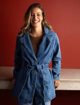 Chaqueta larga de jean azul con cuello con solapa, abertura central con botones, bolsillos delanteros, cintura con pasadores y lazo de ajuste, y puños con botón.