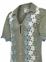 Sweater verde de tejido tipo crochet, con cuello camisero, mangas cortas y cierre frontal de botones. Presenta un diseño de bandas laterales con aplicaciones florales en tonos blancos y celestes.