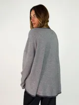Sweater de punto gris con cuello redondo, mangas largas y diseño asimétrico, siendo más corto en la parte delantera y más largo en la trasera, con detalles de terminación en contraste.