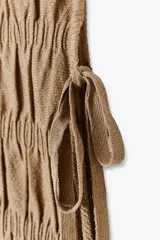 Top de manga larga color beige, con textura fruncida en todo el cuerpo y mangas. Presenta cuello redondo y bajo asimétrico con aberturas laterales ajustables con cordones.