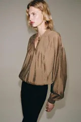 Camisa fluida de Zara Woman Collection, confeccionada en hilatura con mezcla de viscosa en tono tostado. Presenta cuello redondo, mangas largas con puños elásticos y diseño fruncido en la parte delantera. Dispone de cierre frontal con botones ocultos por solapa.