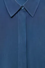 Camisa de corte fluido con cuello solapa y manga corta con dobladillo. Presenta un cierre frontal con botones ocultos bajo una tapeta y un diseño con bajo ligeramente más largo en la parte posterior.