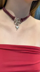 Gargantilla tipo choker confeccionada en tiras de gamuza color bordó, con herrajes metálicos plateados y un dije central en forma de corazón con diseño de efecto derretido y la palabra LOVE grabada.
