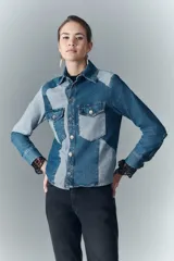 Campera recta de denim rígido con combinación de recortes en tonos de azul. Cuenta con bolsillos de solapa en el pecho y botones metálicos. Las mangas tienen puños con encaje negro.