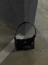 Bolso de hombro negro de charol, con forma alargada y cierre principal con cremallera. Presenta un dije metálico con forma de estrella de cuatro puntas en el frente y detalles de cremalleras decorativas en los laterales.