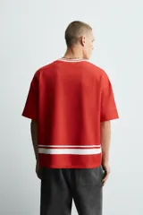Camiseta de manga corta color rojo, corte boxy fit, con cuello pico ribeteado en blanco. Presenta un estampado frontal con el texto "Social Leagues", una estrella blanca y un escudo con el número "4". Tiene detalles de franjas blancas en el bajo y en los hombros.
