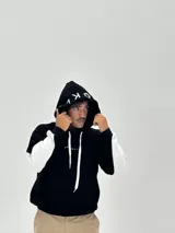 Canguro con capucha de corte boxy, color negro con una franja horizontal blanca que recorre la espalda y las mangas, presentando la palabra 'BROOKLYN' estampada en negro sobre la franja. La capucha también incluye detalles estampados.