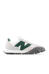 Championes New Balance de estilo casual, con capellada en gamuza color gris claro, logo "N" en verde oscuro y suela de goma con diseño texturizado en gris oscuro.