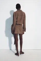 Vestido corto marrón con rayas verticales blancas, cuello camisero, manga larga y detalle de nudo y lazada en la falda. Cuenta con cierre frontal de botones.