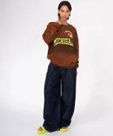 Buzo de algodón y poliéster, color azul oscuro con efecto desgastado/tie-dye. Presenta un gran estampado frontal con la palabra "MICHIGAN" en amarillo con borde negro, un corazón naranja y apliques de pines decorativos en la parte superior. Tiene cuello redondo con terminación deshilachada y bolsillo canguro frontal.