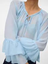 Blusa de organza color celeste, con cuello en V, cierre frontal con tiras y mangas largas con volados. Presenta un diseño traslúcido y holgado.