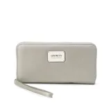 Billetera larga con cierre perimetral, confeccionada en material sintético texturizado color mostaza. Cuenta con correa de mano extraíble y placa metálica con logo de la marca en el frente.