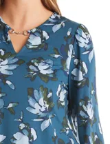 Blusa de seda fría con estampado floral en tonos verdes y azules. Presenta escote en V con un aplique metálico decorativo, mangas largas con puños y corte recto.