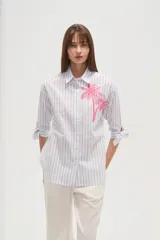 Camisa de algodón con rayas verticales azules y blancas, corte ancho y manga larga con lazos ajustables en los puños. Presenta un bordado de dos palmeras en color rosa brillante en el área del pecho.