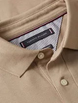 Chomba color beige de Tommy Hilfiger, modelo 1985, de corte regular y mangas cortas. Tiene cuello tipo chomba, cartera de dos botones y logo de la marca bordado en el pecho.