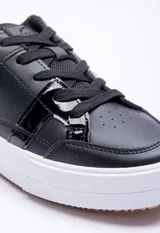Zapatillas urbanas negras con detalle charol en el lateral y suela blanca.
