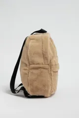 Mochila de textura suave tipo sherpa en color beige, con compartimento principal y bolsillo frontal con cierre. Incluye correas negras ajustables.