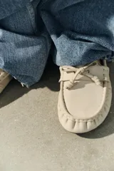 Mocasín plano de piel color beige, con diseño fruncido en la parte delantera, detalle de lazo frontal y cordón lateral entrelazado con ojalillos metálicos.