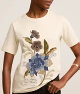 Remera de algodón color beige con cuello redondo y mangas cortas. Presenta un estampado frontal con diseño floral de estilo mosaico y texto en la parte inferior.