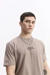 Remera de manga corta color beige con cuello redondo y estampado grande en la espalda. El estampado incluye un gráfico abstracto en tonos naranjas y morados, la palabra "Persistence" en vertical y un texto motivacional debajo.