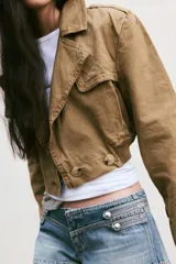 Trench corto de cuello solapa y manga larga, con diseño cruzado y cierre frontal con botones. Presenta bajo acabado en elástico y trabillas en los hombros.