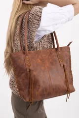 Cartera tipo tote bag de cuero vacuno marrón, con detalles de trenzado artesanal en los laterales y flecos colgantes decorativos. Cuenta con doble asa de hombro y cierre superior.