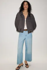 Jeans de tiro alto con pernera ancha y corte cropped. Presenta diseño de cinco bolsillos, cierre frontal con cremallera y botón, e incluye un cinturón de gamuza marrón con hebilla metálica.