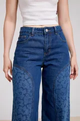 Pantalón de jean color marrón con diseño de paneles laterales estampados con motivos florales. Presenta un corte recto y holgado, con tiro medio y cierre frontal.