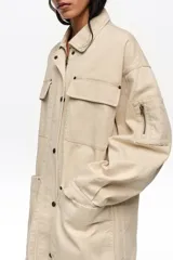 Chaquetón de corte recto color beige, confeccionado en algodón, con cierre frontal de cremallera oculta y botones a presión. Posee cuello camisero, mangas largas, bolsillos con solapa en el pecho, bolsillos tipo plastrón en la parte inferior y un pequeño bolsillo con cierre en la manga.