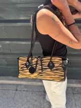 Cartera tipo baguette con estampado animal print de tigre en tonos beige y negro. Tiene detalles en cuero negro, doble cierre superior y correa de mano.