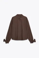 Camisa marrón de manga larga con cuello solapa y cierre frontal de botones. Los puños terminan con lazos anudados.