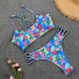 Bikini de dos piezas con estampado tropical de flores y hojas en tonos rosados, azules y verdes sobre fondo celeste. El corpiño es de diseño triangular con espalda cruzada, y la bombacha es cola less con tiras laterales decorativas.