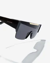 Lentes de sol Hawkers unisex, modelo Paula Echevarría Rubin, con montura negra y lente espejada gris con detalle dorado en la parte superior.
