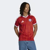 Camiseta de fútbol Adidas de la Selección Chilena, modelo local 26, color rojo con patrón de chevrons en relieve. Presenta cuello en V con ribete blanco y azul, y ribetes blancos en las mangas. Incluye el escudo de la federación chilena y el logo de Adidas en blanco.