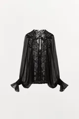Blusa de georgette negro con mangas largas abullonadas, puños con volados y cierre frontal con lazada. Presenta una delicada aplicación de encaje floral a tono en el frente y el cuello.