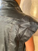 Chaleco sin mangas de gabardina con acabado foil (efecto satinado/brillante), color gris oscuro o negro. Presenta cuello alto y pespuntes contrastantes visibles en el frente y hombros.