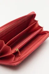 Billetera de formato rectangular con cierre perimetral, confeccionada en material símil cuero de textura suave. Presenta un aplique metálico circular con logo grabado en el frente y tirador de cierre en el mismo material.