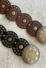 Cinturón ancho de cuero negro con diseño de círculos superpuestos. Presenta apliques metálicos circulares en tono dorado envejecido, algunos lisos y otros con grabado tribal/étnico. Los círculos de cuero tienen ojales metálicos pequeños alrededor de los apliques grandes.