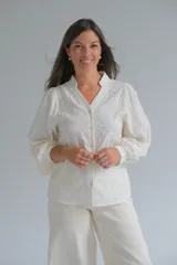 Blusa de color crudo con cuello mao y cierre frontal de botones. Presenta mangas largas abullonadas con puños y un delicado diseño bordado con relieve en toda la prenda.