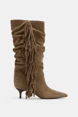 Bota de caña alta confeccionada en serraje, con diseño fruncido y detalle de flecos laterales. Presenta tacón kitten y acabado en punta.