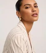 Blazer de poliéster color crudo con rayas verticales finas en marrón. Tiene cuello con solapas peaked, bolsillos delanteros y mangas largas con botón para arremangar. Sin forro.