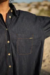 Vestido camisero corto de jean azul oscuro, con cuello clásico, mangas 3/4 con puños arremangados y cierre frontal con botones dorados.