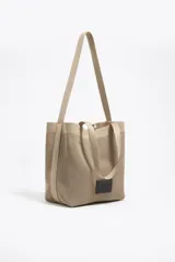 Bolso shopper mediano de tejido de punto color beige, con doble asa corta de mano y una correa larga ajustable para el hombro. Incluye parche con logo en la parte frontal.