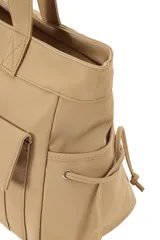 Cartera tipo shopper de poliuretano color beige, con dos bolsillos frontales con solapa, tirantes largos para el hombro y cordones ajustables en los laterales.