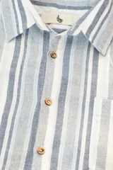 Camisa de manga corta con rayas verticales en tonos azul, gris y blanco. Tiene cuello clásico, botones de coco y un bolsillo en el pecho.