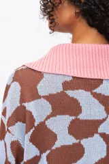 Buzo de punto con diseño geométrico abstracto en tonos marrón y celeste. Presenta cuello tipo polo en color rosado con cierre frontal de botones, mangas largas con puños rosados y un pequeño parche con logo en la manga izquierda.
